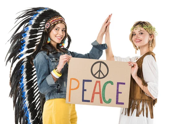 Dos chicas hippies bisexuales sonrientes en tocado indio y guirnalda sosteniendo pancarta con inscripción y tocando manos aisladas en blanco - foto de stock