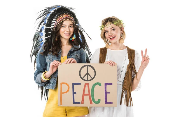 Deux hippies bisexuelles en coiffe indienne et couronne tenant une pancarte avec inscription et montrant un signe de paix isolé sur blanc — Photo de stock