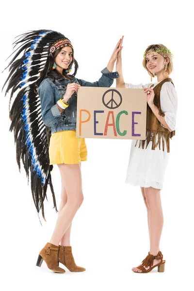 Visão comprimento total de duas meninas hippies bissexuais em cobertura para a cabeça indiana e coroa segurando cartaz com inscrição e tocar as mãos isoladas no branco — Fotografia de Stock