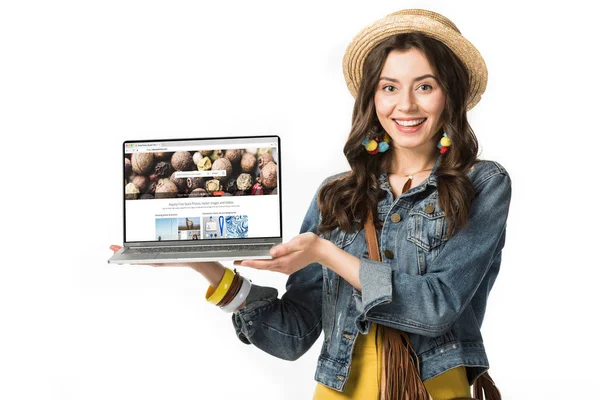 KYIV, UCRAINA - 4 FEBBRAIO 2019: ragazza hippie sorridente in cappello di paglia che tiene il computer portatile con sito web di foto di deposito sullo schermo isolato su bianco — Foto stock