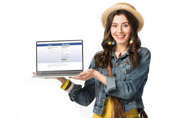 QUIIV, UCRÂNIA - FEVEREIRO 4, 2019: menina hippie sorridente em chapéu de palha segurando laptop com site facebook na tela isolada no branco — Fotografia de Stock