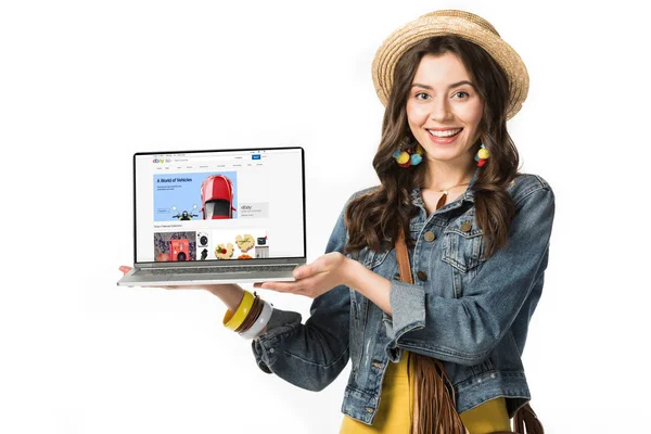 KYIV, UCRANIA - 4 DE FEBRERO DE 2019: niña hippie sonriente en sombrero de paja que sostiene el ordenador portátil con el sitio web de ebay en la pantalla aislada en blanco - foto de stock