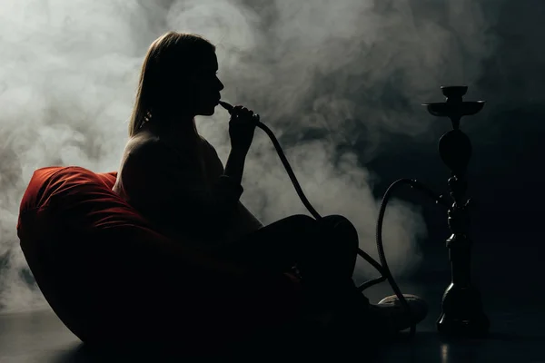 Silueta de la mujer sentada en la silla de la bolsa de frijol y fumar narguile en la oscuridad - foto de stock