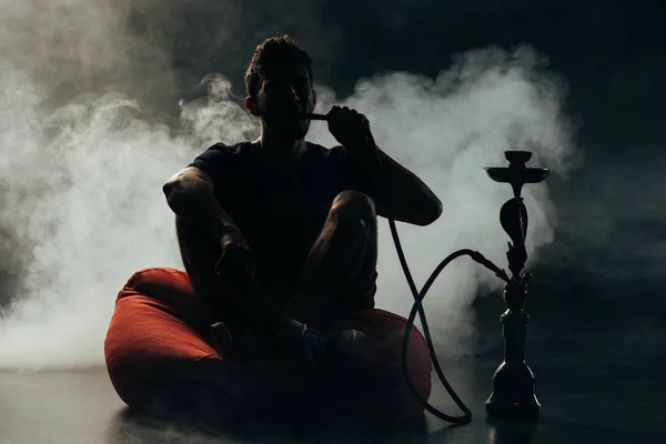 Silhouette of man sitting on bean bag chair and smoking hookah in darkness — Stock Photo