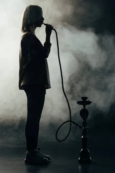Vista lateral de la silueta de la mujer fumar narguile en la oscuridad - foto de stock