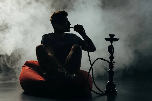 Silhouette of man sitting on bean bag chair  with crossed legs and smoking hookah — Stock Photo