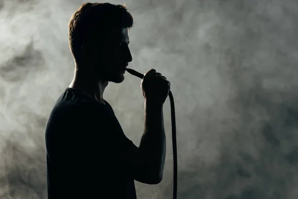 Silhouette of man smoking hookah in smoke in darkness — Stock Photo