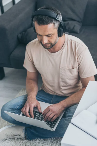 Schöner Mann mit Kopfhörer, der Laptop benutzt, während er auf dem Boden sitzt — Stockfoto