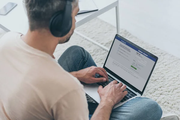 KYIV, UKRAINE - 5 MAI 2019 : Homme dans un casque assis sur le sol et utilisant un ordinateur portable avec application Facebook à l'écran . — Photo de stock