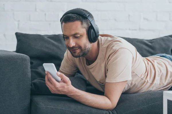 Un homme souriant dans un casque couché sur un canapé et utilisant un smartphone — Photo de stock