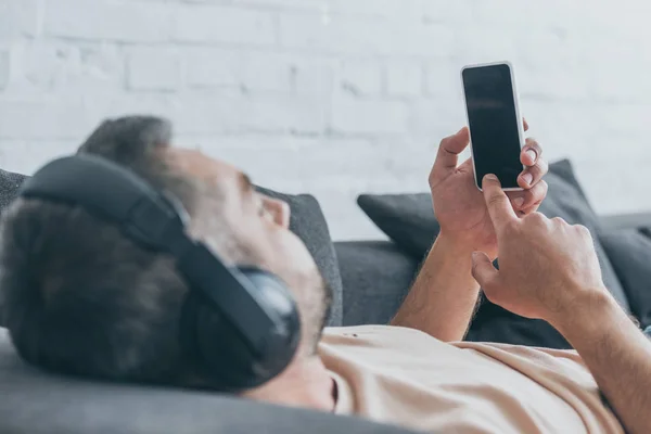 Enfoque selectivo del hombre en auriculares con teléfono inteligente con pantalla en blanco mientras descansa en el sofá - foto de stock