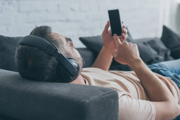 Hombre adulto en auriculares tumbado en el sofá y la celebración de teléfono inteligente con pantalla en blanco - foto de stock