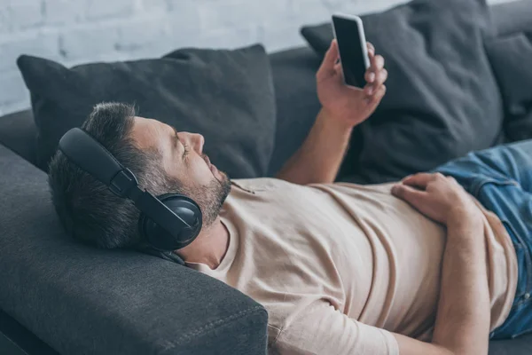 Homem adulto em fones de ouvido segurando smartphone com tela em branco enquanto descansa no sofá — Fotografia de Stock