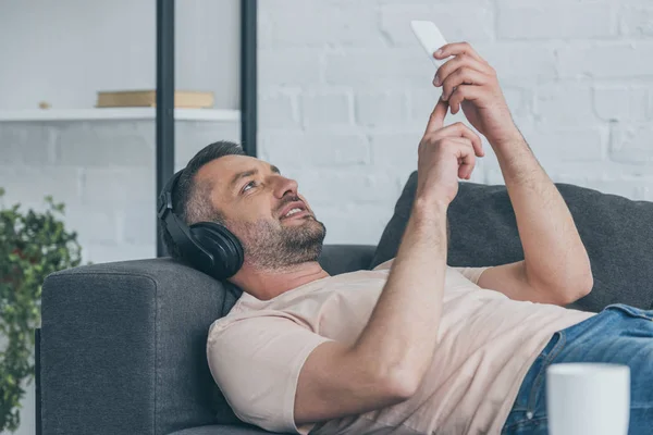 Smiling man in headphones lying on sofa and using smartphone — Stock Photo