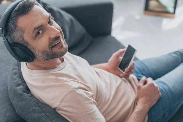 Bel homme dans les écouteurs souriant à la caméra tout en étant assis sur le canapé et tenant smartphone avec écran blanc — Photo de stock
