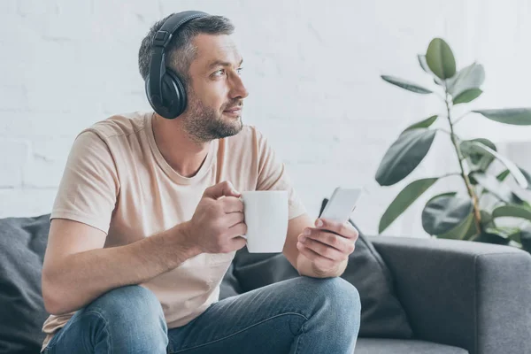 Homem bonito em fones de ouvido olhando embora enquanto segurando xícara de café — Fotografia de Stock