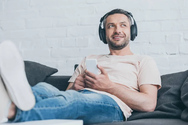 Foco seletivo do homem alegre em fones de ouvido olhando embora enquanto segurando smartphone — Fotografia de Stock