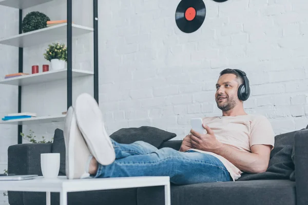 Homme souriant dans les écouteurs assis avec les jambes croisées sur la table et tenant smartphone — Photo de stock