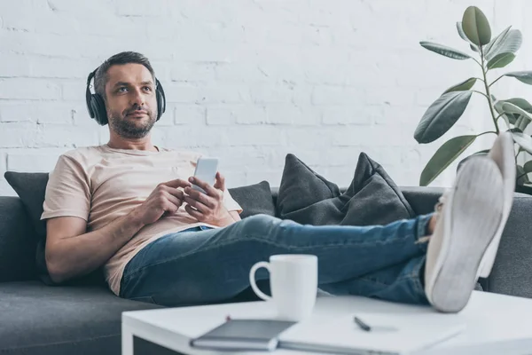 Selektiver Fokus eines gutaussehenden Mannes mit Kopfhörern, der sein Smartphone in der Hand hält und wegschaut, während er mit überkreuzten Beinen auf dem Tisch sitzt — Stockfoto