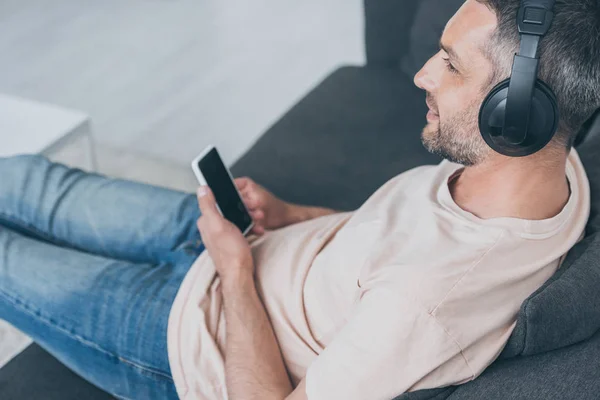 Homem adulto em fones de ouvido segurando smartphone com tela em branco enquanto sentado no sofá — Fotografia de Stock