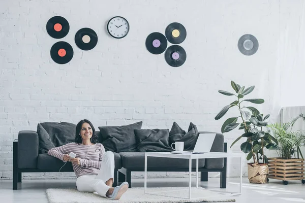 Fröhliche Frau, die Musik im Kopfhörer hört und das Smartphone hält, während sie zu Hause auf dem Boden sitzt — Stockfoto