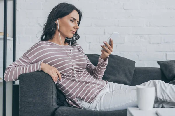 Mujer pensativa sentada en el sofá, sosteniendo el teléfono inteligente y escuchando música en los auriculares - foto de stock