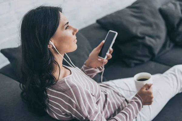 Вдумчивая молодая женщина держит смартфон с чистым экраном и смотрит в сторону, слушая музыку в наушниках — стоковое фото