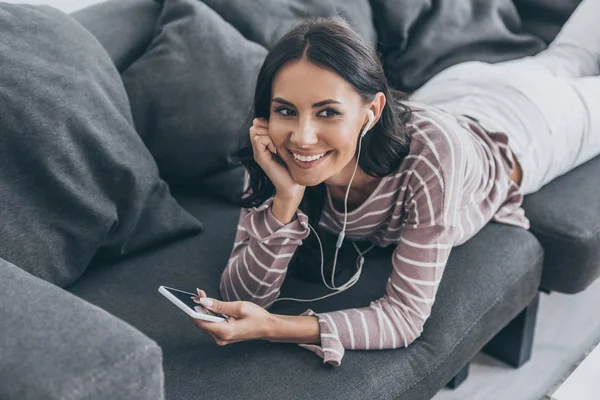 Mulher alegre olhando embora enquanto descansa no sofá com smartphone e ouvir música em fones de ouvido — Fotografia de Stock