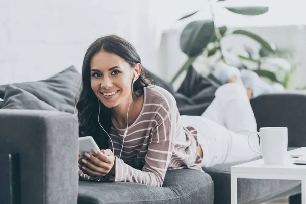 Mujer sonriente mirando a la cámara mientras está acostada en el sofá y escuchando música en los auriculares - foto de stock