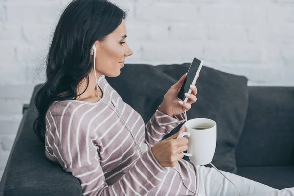Femme souriante utilisant un smartphone tout en écoutant de la musique dans les écouteurs et en tenant une tasse de café — Photo de stock