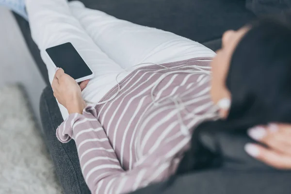 Enfoque selectivo de la mujer joven acostada en el sofá y la celebración de teléfono inteligente con pantalla en blanco - foto de stock