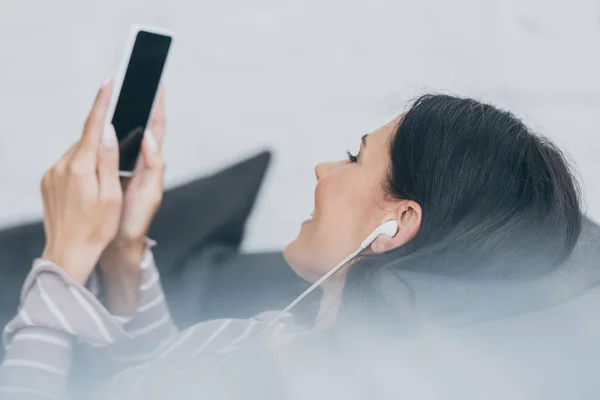 Foyer sélectif de la jeune femme écoutant de la musique dans les écouteurs tout en étant couché sur le canapé et tenant smartphone avec écran blanc — Photo de stock