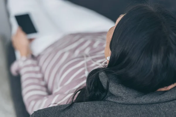 Вид зверху молодої жінки, що лежить на дивані, слухає музику в навушниках і тримає смартфон — стокове фото