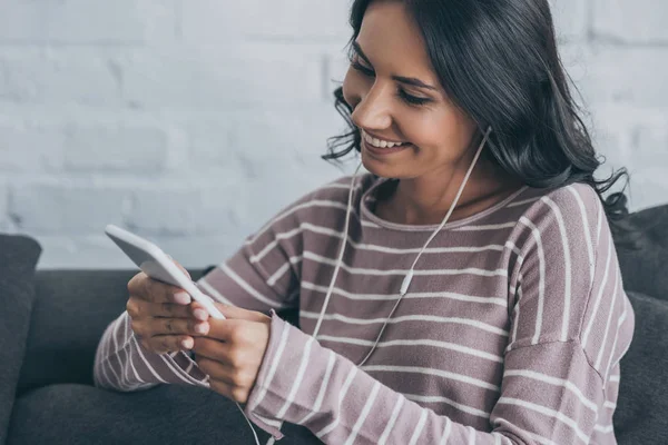 Mujer alegre usando el teléfono inteligente mientras está sentado en el sofá y escuchar música en los auriculares - foto de stock