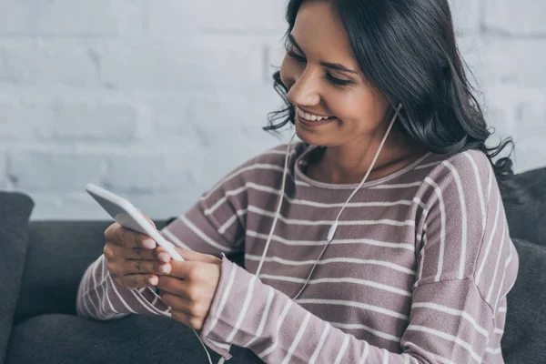 Donna allegra utilizzando smartphone mentre seduto sul divano e ascoltare musica in auricolare — Foto stock