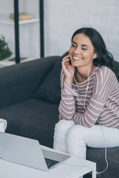 Happy woman with closed eyes listening music in earphones while sitting near desk with laptop — Stock Photo