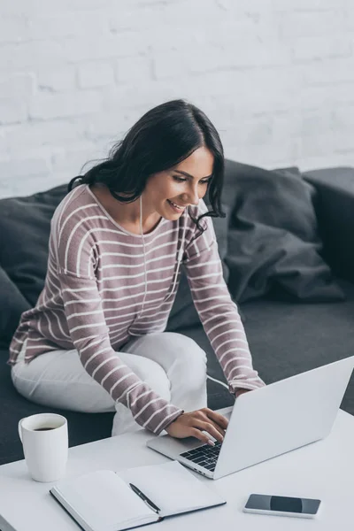 Femme gaie écouter de la musique dans les écouteurs tout en étant assis au bureau et en utilisant un ordinateur portable — Photo de stock