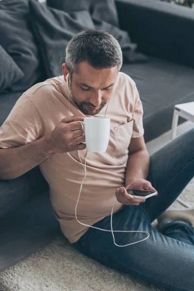 Homem bonito bebendo café enquanto ouve música em fones de ouvido e usando smartphone — Fotografia de Stock