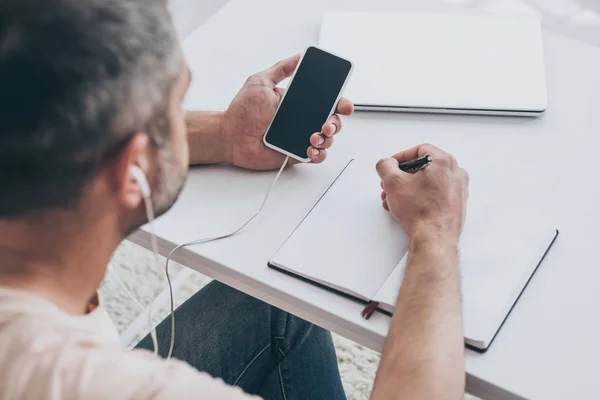 Overhead view of man in earphones holding smartphone with blank screen and writing in notebook — Stock Photo