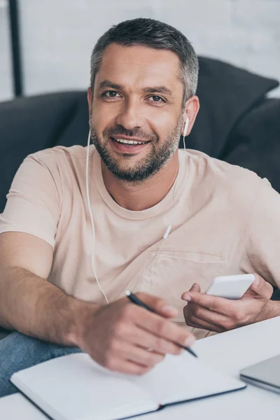 Hombre guapo mirando a la cámara mientras escucha música en los auriculares y sosteniendo el teléfono inteligente - foto de stock
