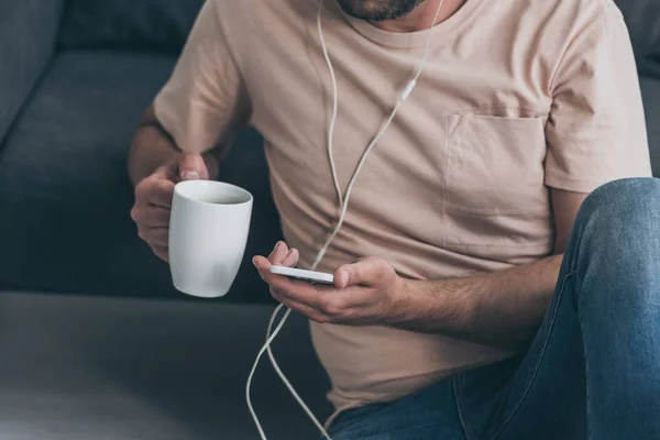 Vista ritagliato di uomo utilizzando smartphone durante l'ascolto di musica in cuffia e tenendo tazza di caffè — Foto stock