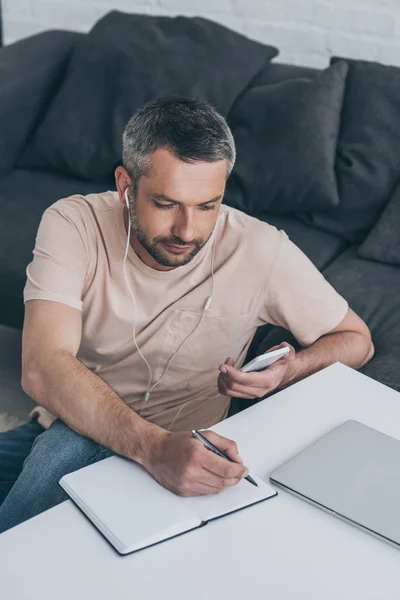 Aufmerksamer Mann mit Kopfhörer sitzt am Tisch, benutzt Smartphone und schreibt in Notizbuch — Stockfoto