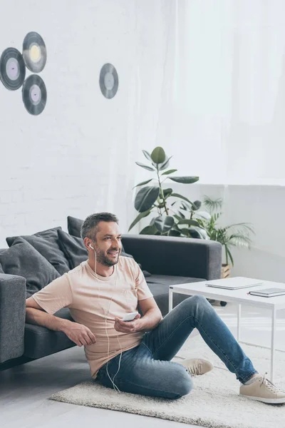 Cheerful man sitting on floor, listening music in earphones and looking away — Stock Photo