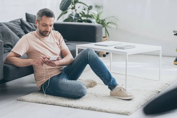 Handsome man sitting on floor at home, using smartphone and listening music in earphones — Stock Photo