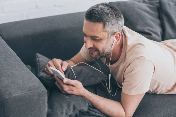 Bel homme allongé sur le canapé, écoutant de la musique dans les écouteurs et utilisant un smartphone — Photo de stock