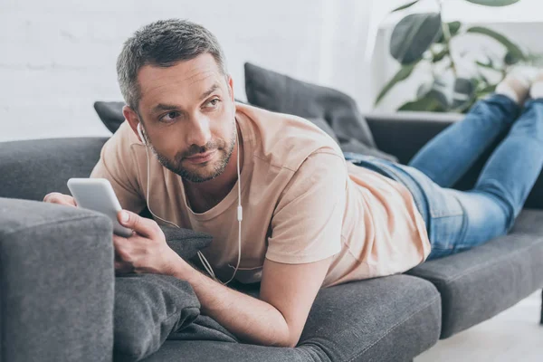 Thoughtful man lying on sofa, listening music in earphones and looking away — Stock Photo