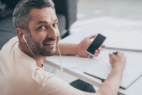 Messa a fuoco selettiva di uomo allegro in cuffia sorridente alla fotocamera mentre tiene smartphone con schermo bianco e scrittura in notebook — Foto stock