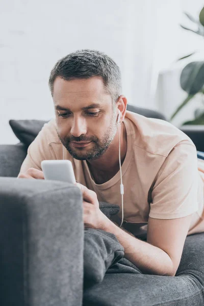 Hombre guapo acostado en el sofá, escuchando música en los auriculares y utilizando el teléfono inteligente - foto de stock