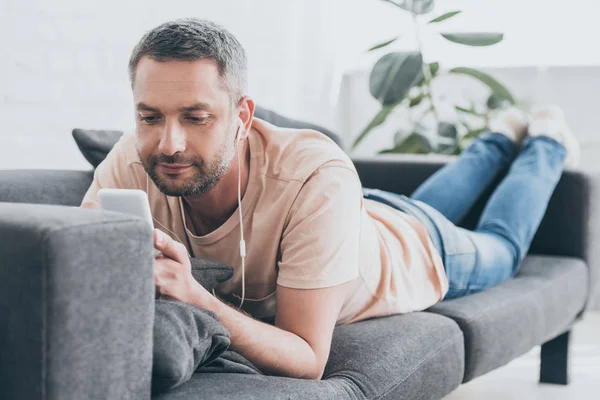 Усміхнений чоловік слухає музику в навушниках і використовує смартфон під час відпочинку на дивані — стокове фото