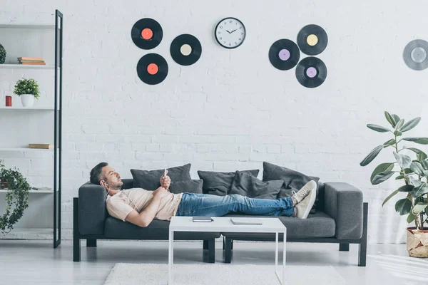 Adult man in earphones using smartphone while lying on sofa in spacious living room — Stock Photo